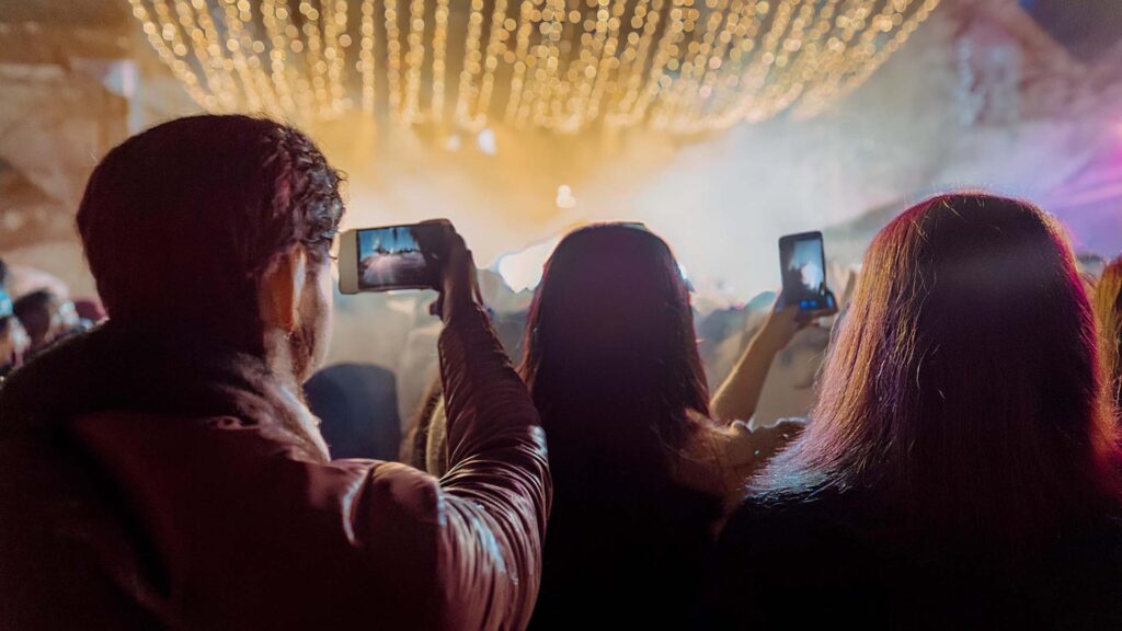 مجموعة من الأشخاص وسط احتفال ليلي يظهرون من الخلف، يحملون هواتف وكاميرات يصوّرون شيئاً ما